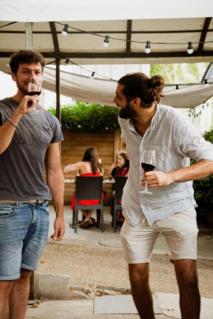 Deux hommes souriants boivent du vin rouge sur une terrasse éclairée, l'un regardant l'objectif, l'autre riant en discutant.