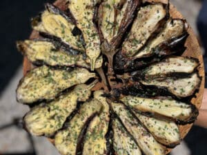Aubergines rôties coupées en deux, nappées de sauce crémeuse, ciboulette fraîche et sumac. Disposées en cercle sur planche en bois.