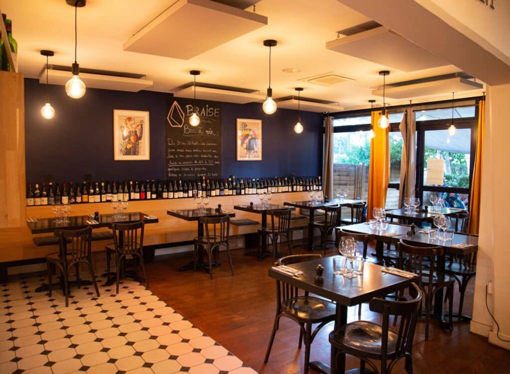 Intérieur élégant de Braise Bar à vin : murs bleus, bouteilles, tables en bois foncé, chaises, sol carrelé, lumières chaudes. Ambiance conviviale.