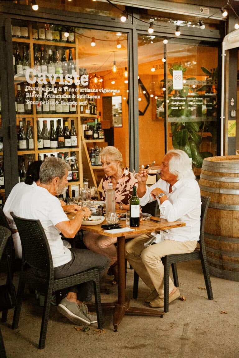 Des convives dînent sur une terrasse devant un magasin Caviste offrant Champagnes, Vins d'artisans, Bières et Spiritueux.