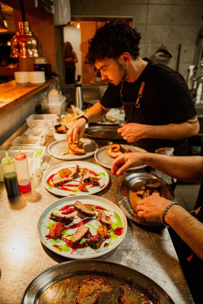 Chef talentueux en pleine préparation de plats gastronomiques Chef en cuisine préparant plusieurs plats gastronomiques. Plats de viande et poulpe sur comptoir métallique. Mains expertes dressant les assiettes.