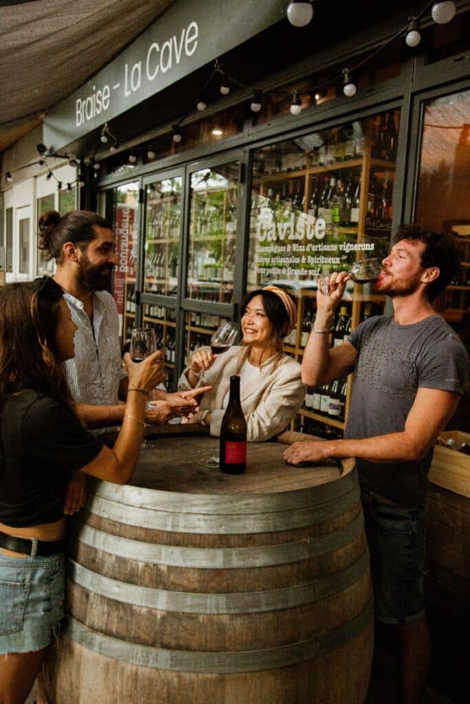 Amis dégustant du vin autour d'un tonneau devant le caviste 'Braise - La Cave'. Ambiance conviviale.