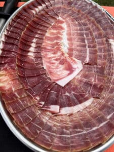 Grand plat argenté rempli de tranches fines de jambon ibérique salé, arrangées en éventail avec du gras persillé.