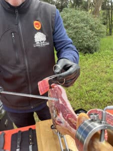 Professionnel ganté découpe du jambon Noir de Bigorre sur support, tenant une tranche fine avec pince. Le logo "LE NOIR DE BIGORRE" est visible sur son gilet.