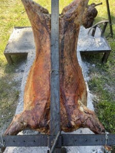 Agneau ou chevreuil entier rôti à la broche, peau croustillante et dorée, cuisant lentement sur un lit de braises en extérieur. Un rôti ficelé est aussi suspendu.