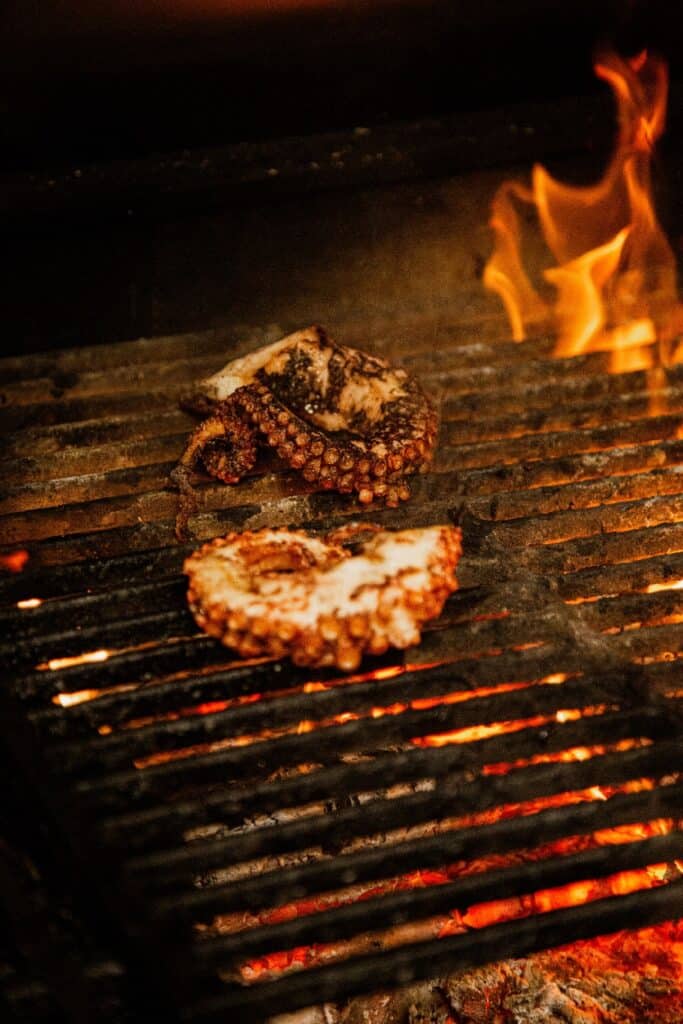 Poulpe grillé au barbecue : cuisson parfaite et flammes Deux morceaux de poulpe grillent sur un barbecue avec des flammes vives à droite et des braises rouges sous la grille.