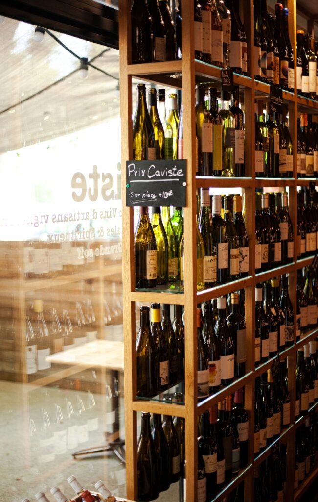 Étagères en bois remplies de bouteilles de vin variées dans une cave. Un panneau indique 'Prix Caviste, Sur place +10€'.