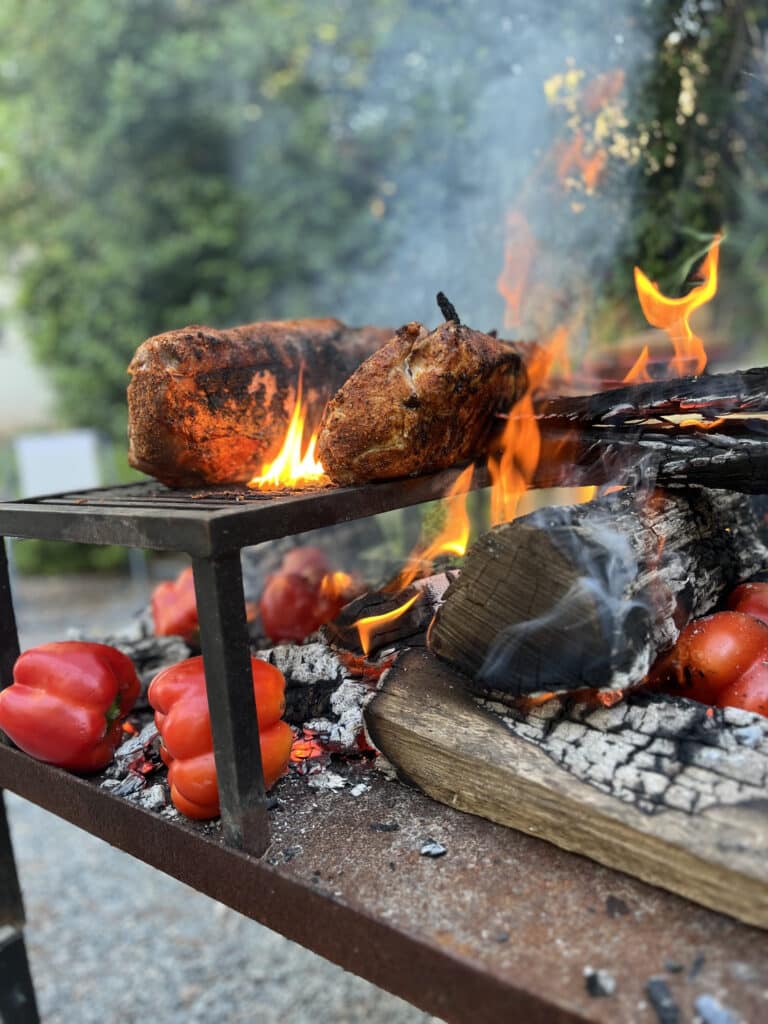 Deux morceaux de viande rôtissant sur un gril en métal au-dessus de flammes intenses. Des poivrons rouges grillent sur les braises d'un barbecue extérieur.